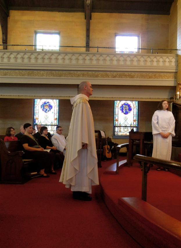 Eine Frau und ein Mann in weißen Kleidern stehen in der Mitte eines Raums mit rotem Teppich, hölzernen Geländern und Musikinstrumenten, mit Menschen, die links auf Bänken sitzen und Fenstern im Hintergrund.