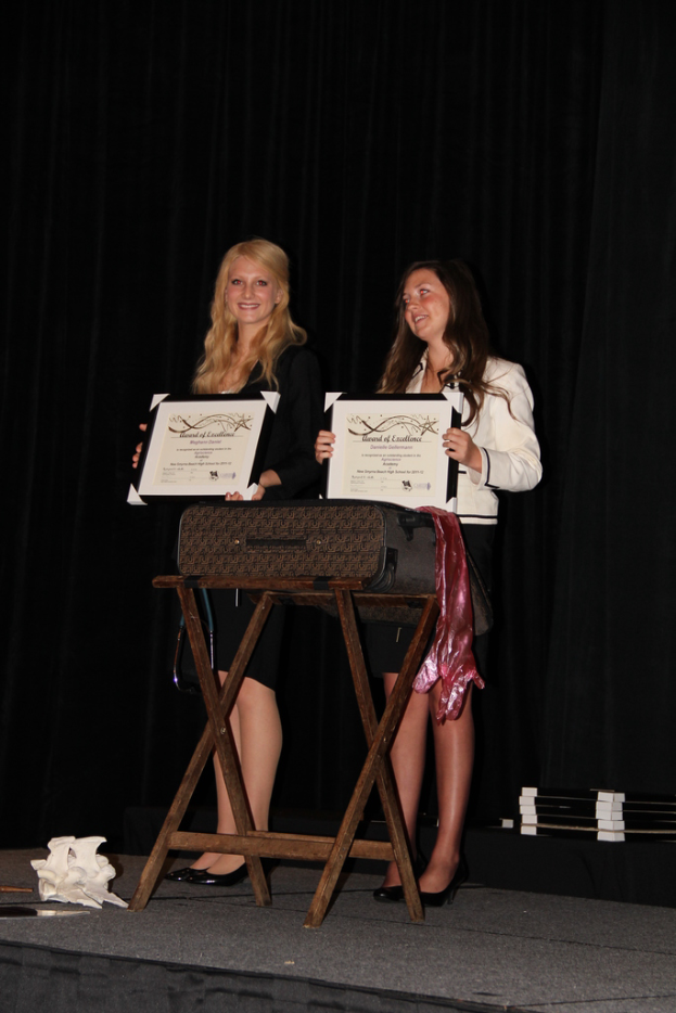 Zwei Frauen, die strahlen und Zertifikate halten, vor einem Tisch mit einer Schachtel darauf, vor einem schwarzen Vorhang.