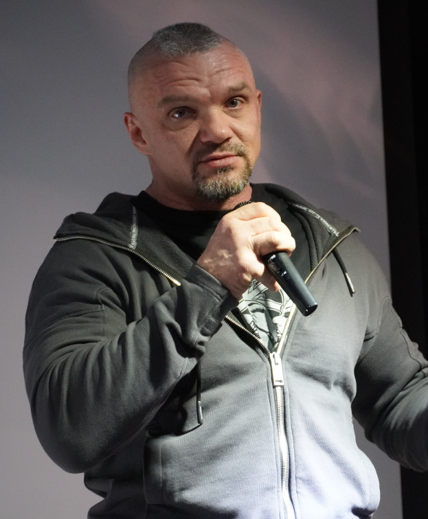 Ein Mann in schwarzer Kapuzenjacke, schwarzem Anzug und weißem Hemd hält ein Mikrofon und steht selbstbewusst vor einer Wand.
