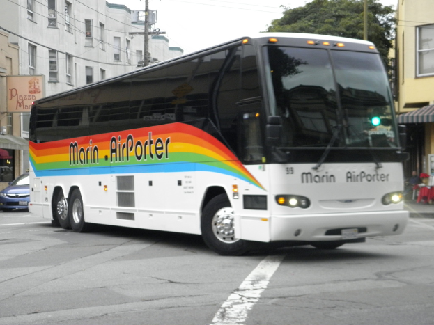 Ein Bus fährt auf einer Straße mit Gebäuden, Bäumen und Masten im Hintergrund.
