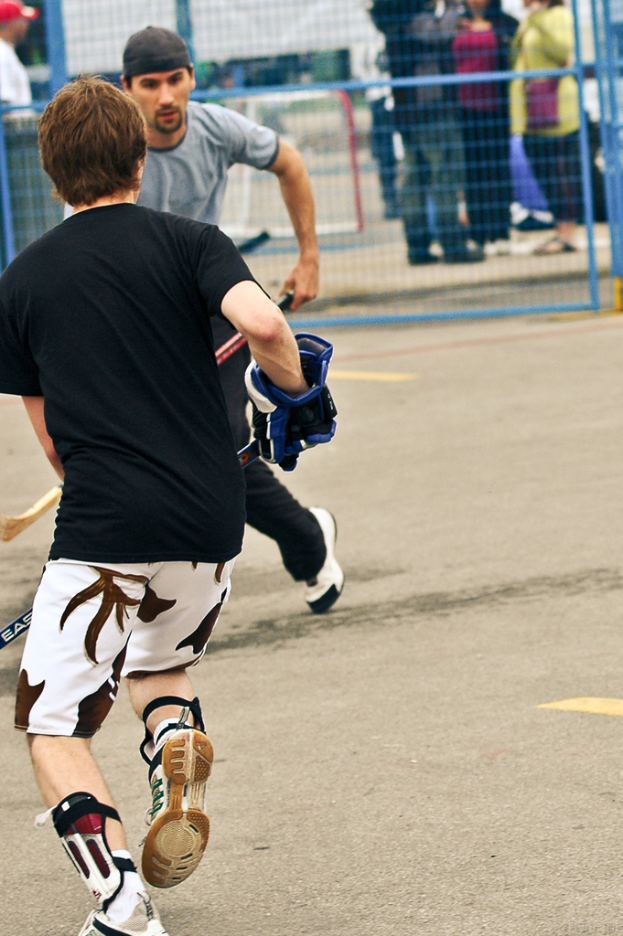 Zwei Personen spielen Hockey auf einer Straße mit einem Zaun im Hintergrund und einer Zuschauermenge im Hintergrund.