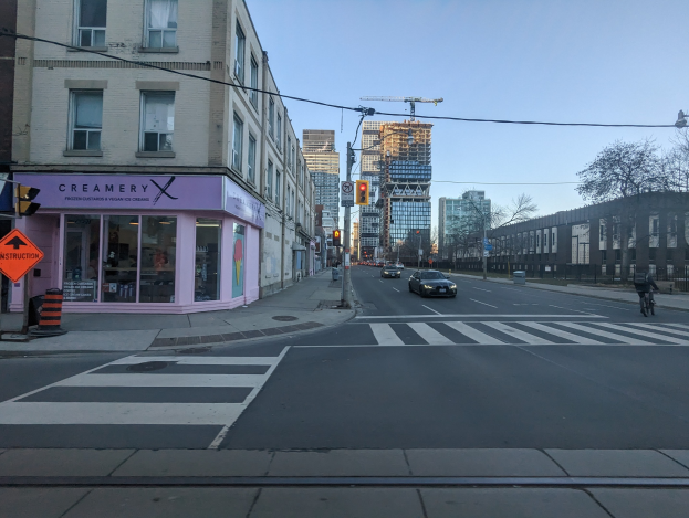 Stadtstraße mit fahrenden Autos, ein Radfahrer auf dem Gehweg, Verkehrsampeln, Schilder, Gebäude, Bäume und Himmel.