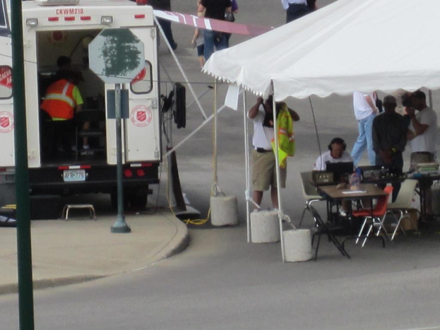 Ein Rettungswagen befindet sich auf der linken Seite einer Straße, während auf der rechten Seite ein Zelt eine Gruppe von Menschen schützt, mit einem Tisch und einem Stuhl in der Nähe.