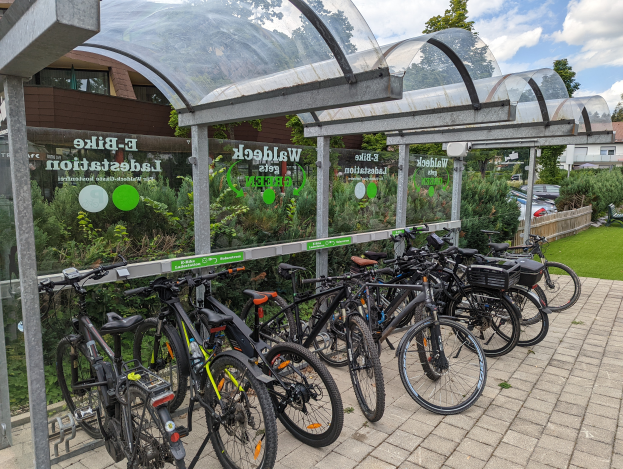 Eine Reihe von Fahrrädern, die vor einem glaswandigen Gebäude mit einem Schuppen, Bäumen, Pflanzen, Fahrzeugen auf der Straße und einem bewölkten Himmel geparkt sind, wahrscheinlich Teil eines E-Bike-Netzwerks in den Niederlanden.