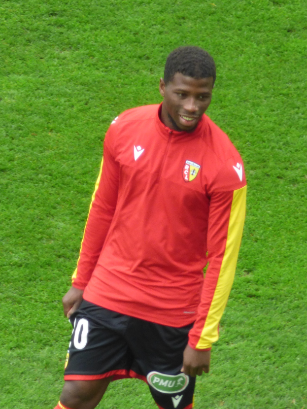 Ein Mann in rotem Trikot und schwarzen Shorts steht auf einem Fußballfeld, lachend, bereit zu spielen als Flügelspieler.