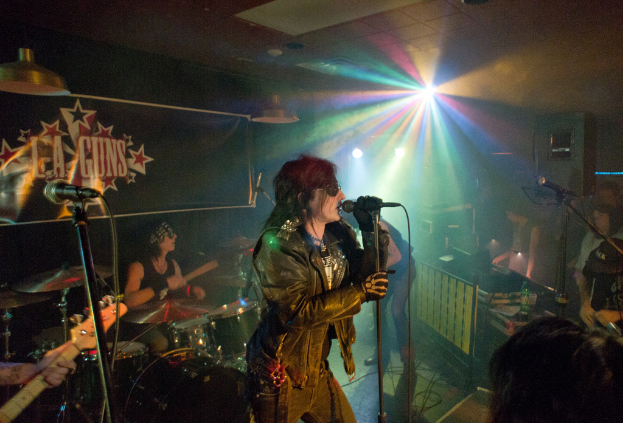 Eine Frau in einer schwarzen Jacke singt in ein Mikrofon auf der Bühne, mit einem Mann in Sonnenbrille und Stirnbinde, der hinter ihr ein Instrument spielt, unter einem Schild, auf dem "LA Guns" steht und bunte Diskoleuchten.