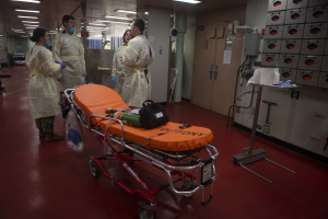 Gruppe von Menschen in Schutzausrüstung um eine Trage in einem Krankenhauszimmer mit medizinischer Ausrüstung, darunter ein Bett, ein Ständer, eine Tür, eine Säule, ein Schild und Deckenleuchten.