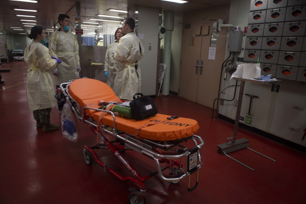 Gruppe von Menschen in Schutzausrüstung um eine Trage in einem Krankenhauszimmer mit medizinischer Ausrüstung, darunter ein Bett, ein Ständer, eine Tür, eine Säule, ein Schild und Deckenleuchten.