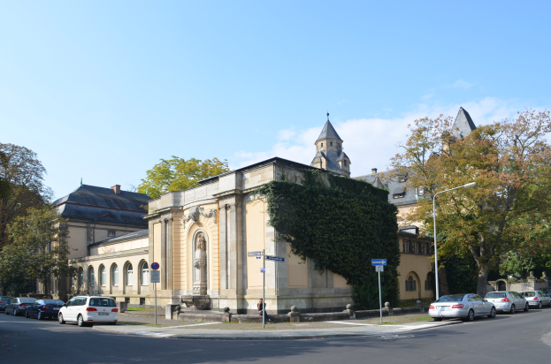 Die Kirche St. Peter und St. Paul in Heidelberg, Deutschland, ein großes Gebäude mit einem zentralen Uhrenturm, umgeben von Bäumen, Straßenlaternen, Straßenlaternen, Kraftfahrzeugen, Fußgängern und Verkehrszeichen, unter einem bewölkten Himmel.