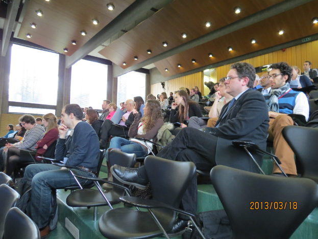 Eine Gruppe von Menschen, die in einem Konferenzsaal mit einem geschlossenen Glasfenster und einem Dach mit ein paar Lampen darüber sitzen.