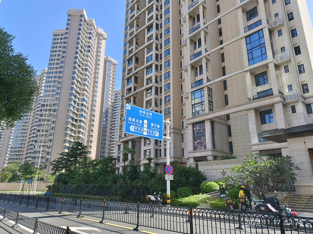 Eine Straße in einer Stadt in Hong Kong mit hohen Wohngebäuden, Bäumen, Pflanzen, einem Zaun, Schildern, Pfählen, Fahrzeugen und einem klaren blauen Himmel.