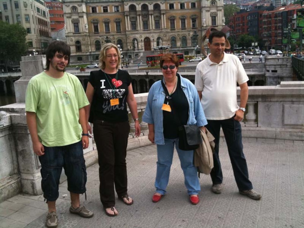 Menschen, die vor einem Geländer und einer Säule stehen und lächeln, mit Fahrzeugen, Gebäuden, Bäumen und anderen Menschen im Hintergrund.