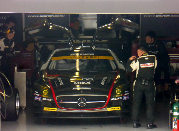 Ein Mercedes-Benz SLS AMG GT3 Rennwagen steht in einer Garage mit zwei Personen davor, einem Feuerlöscher rechts und Tafeln mit Text und Bildern im Hintergrund.