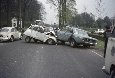 Zwei beschädigte Autos am Straßenrand mit Menschen in der Nähe, vor dem Hintergrund von Bäumen, Strommasten, Gebäuden und einem klaren blauen Himmel.