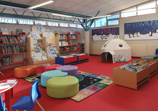 Eine hell erleuchtete Kinderbibliothek mit Bücherregalen, Tischen, Stühlen, Hockern, Teppichen, Wandtafeln, Deckenlampen und großen Fenstern.