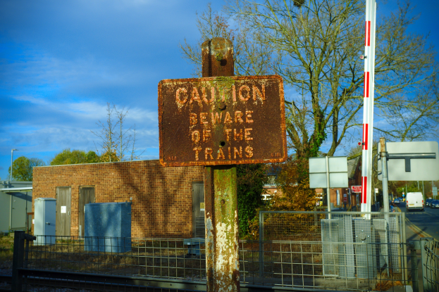 Warnschild an einem Bahnübergang-Zaun mit Bäumen, Masten, einem Gebäude, Containern, Fahrzeugen auf einer Straße und einem bewölktem Himmel.