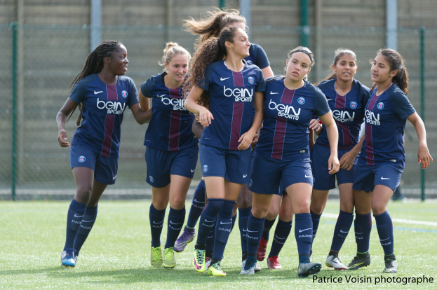 Eine Gruppe junger Frauen beim Fußballspielen auf einem Rasenplatz mit Maschendrahtzaun und einer Wand im Hintergrund, beschriftet mit "Paris Saint-Germain Frauenfußball" in der rechten unteren Ecke.