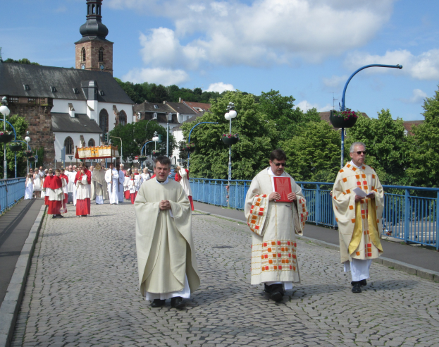 Eine Gruppe von Priestern geht eine Straße entlang, die von Laternenmasten, Geländern und Bäumen gesäumt ist, mit Gebäuden und einem bewölkten Himmel im Hintergrund; ein Priester hält ein Buch.