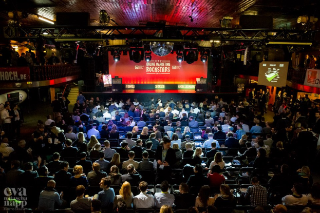 Eine große Menschenmenge sitzt auf Stühlen, die einer Bühne mit Bildschirmen und Bannern zugewandt sind, die von Scheinwerfern beleuchtet und von Geländern umgeben sind, mit einem Wasserzeichen in der linken unteren Ecke, das eine Atmosphäre der Aufregung und Spannung für ein Online-Marketing-Event der Rockstars schafft.