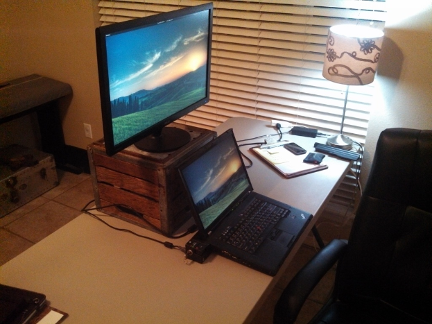 Ein Tisch mit einer Box, Monitor, Laptop, Pad, Papieren, Mobiltelefonen, Laternenlampe und Kabel; ein schwarzer Stuhl; im Hintergrund ein Bett mit einer Box darunter.