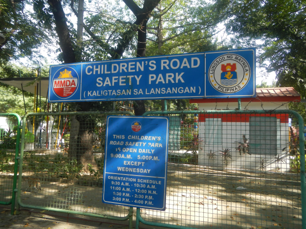 Metallzaun um einen Kinderstraßensicherheitspark mit zwei blauen Informationsschildern, einem kleinen Haus, Pflanzen, Bäumen und einem bewölkten Himmel.