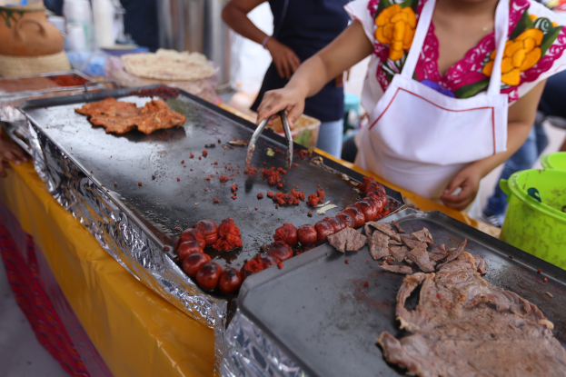Eine Frau in einer Schürze kocht auf einem Grill an einem Markt, mit einem Tisch in der Nähe, auf dem Tabletts mit Lebensmitteln, ein Eimer und andere Gegenstände stehen, während im Hintergrund ein paar Menschen stehen.