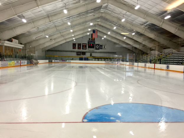 Große Indoor-Eisbahn mit Deckenbeleuchtung, Werbetafeln, Treppen mit Geländern, einer Wand im Hintergrund und einigen Menschen, was auf eine Sportanlage hinweist, die likely für Hockey verwendet wird.