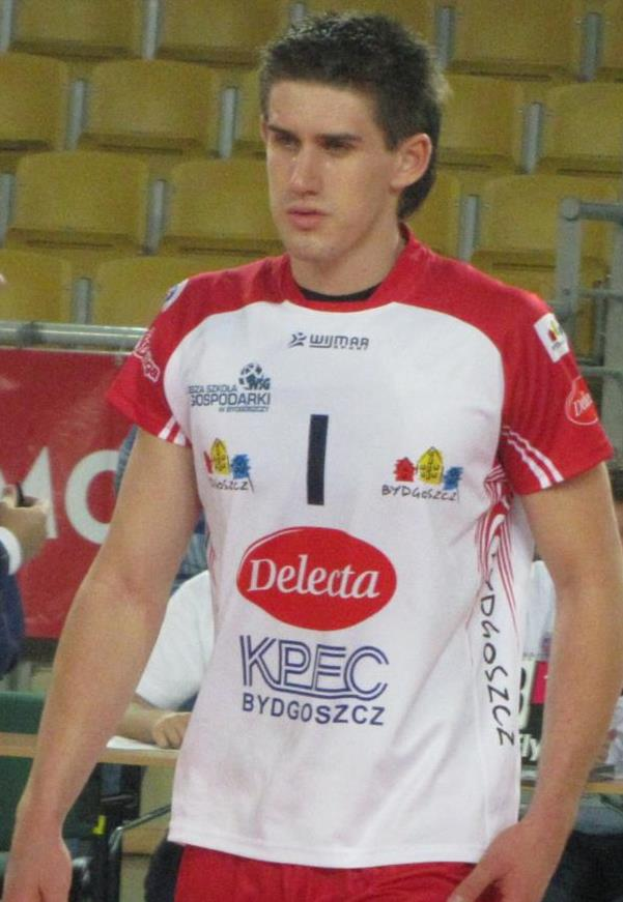 Ein Mann in einem roten und weißen Handballtrikot steht auf einem Feld, umgeben von Zuschauern, mit einem roten Banner im Hintergrund, das 'Krasnoyarsk vs Bydgoszcz' trägt und entschlossen aussieht.