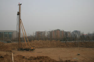 Eine Baustelle mit einem Kran in der Mitte, umgeben von Gebäuden, Bäumen und einem klaren blauen Himmel, mit bodenbedecktem Grund und verstreuten Gegenständen.