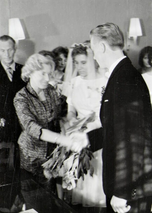 Ein Schwarz-Weiß-Foto einer Braut und eines Bräutigams beim Austausch der Ringe, wobei die Braut ein weißes Kleid und einen Schleier trägt und einen Strauß hält und der Bräutigam einen Anzug und eine Krawatte trägt, mit Wandlampen im Hintergrund.