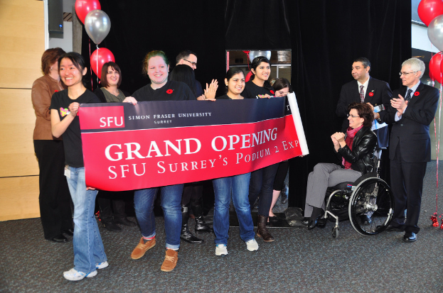 Eine Gruppe von Frauen hält ein Banner hoch, daneben stehen zwei klatschende Männer, eine sitzende Frau im Rollstuhl und ein schwarzer Vorhang mit Ballons im Hintergrund.