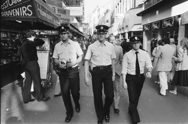 Drei Polizisten in Uniform gehen eine belebte Stadtstraße entlang, gesäumt von hohen Gebäuden und Geschäften, mit Passanten und einem sichtbaren Himmel im Hintergrund.