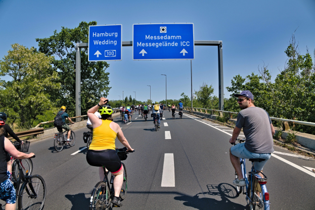 Eine Gruppe von Radfahrern mit Helmen fährt eine Straße mit einer Begrenzung auf einer Seite und Bäumen auf der anderen entlang, unter einem klaren blauen Himmel mit Laternen im Hintergrund; ein Schild oben könnte eine Radtour in Hamburg ankündigen.
