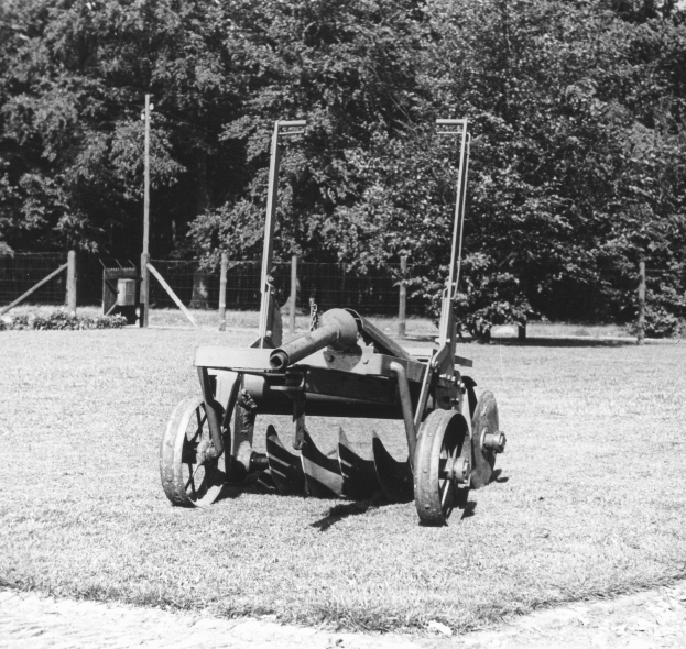 Schwarzes und weißes Foto eines alten Traktors auf einem Feld mit Bäumen, Pfählen und einem Zaun im Hintergrund.
