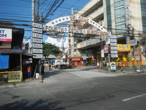 Eine belebte Stadtstraße mit Fahrzeugen, Fußgängern, Gebäuden und einem Schild in der Mitte, unter einem bewölkten Himmel.