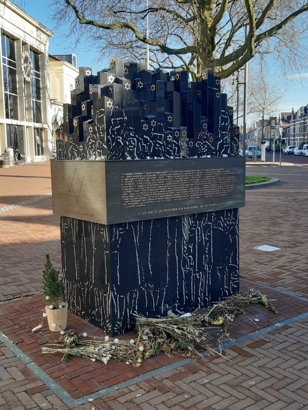 Denkmal in einem Stadtplatz, das an Holocaust-Opfer erinnert, mit einem Baum davor, einem Topf mit einer Pflanze auf dem Boden, verstreuten trockenen Blättern und im Hintergrund Gebäude, Fahrzeuge, Mäste und Bäume.