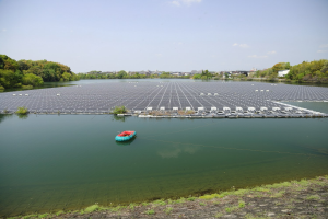 Ein kleines Boot schwimmt auf einem Gewässer, umgeben von üppiger Vegetation und Gebäuden unter einem klaren blauen Himmel, mit Solarpanelen auf der Wasseroberfläche, die auf eine Solar-Farm hinweisen.