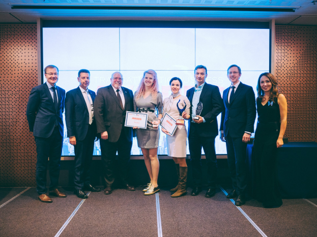 Gruppe von strahlenden Menschen mit Urkunden vor einem Bildschirm mit einem Tisch und Wandobjekt im Hintergrund, Gewinner der 2018 International Business Awards.
