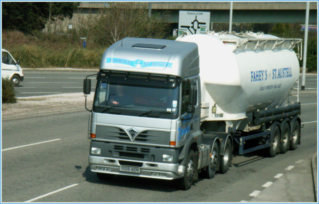 Ein Lkw mit Fahrer √∂ffnet sich auf einer Stra√üe, mit einem Van auf der Gegenfahrbahn, B√§umen und einer Br√ľcke im Hintergrund.
