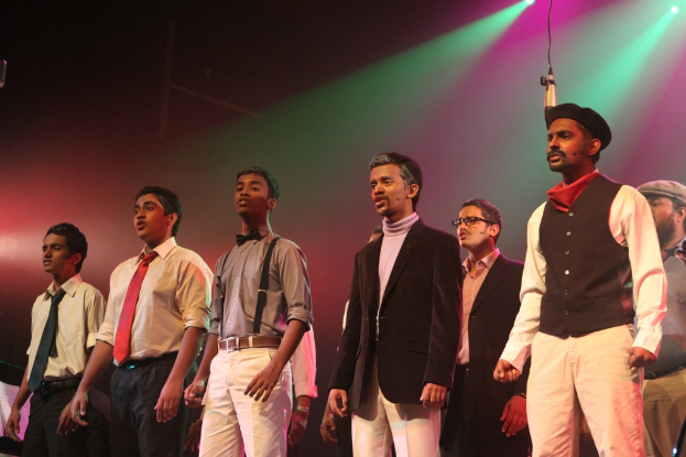 Eine Gruppe von M├Ąnnern steht auf einer B├╝hne und spielt ein Drama, mit gr├╝nen Spotlights dahinter.