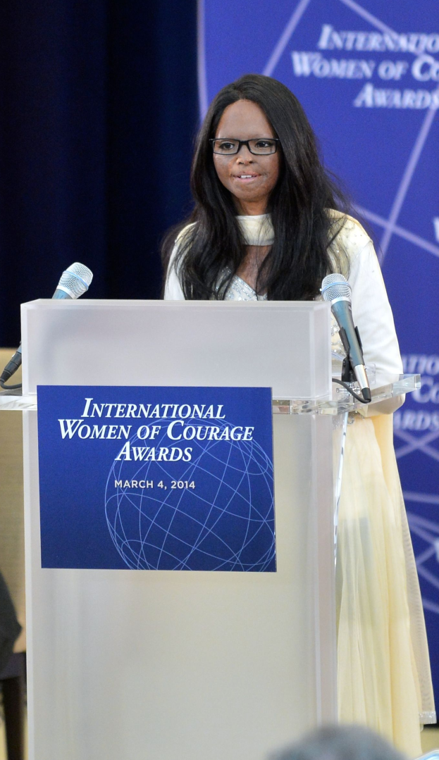 Eine Frau in einem weißen Kleid und Brille steht an einem Podium mit zwei Mikrofonen und einer Tafel, während sie eine Menge auf den "International Women of Courage Awards 2014" anspricht.