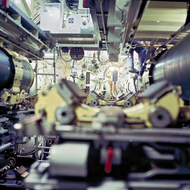 Ein Mann steht auf einer Maschine in einem industriellen Raum, umgeben von anderen Maschinen und scheint Wartungs- oder automatisierungsbezogene Aufgaben durchzuführen.