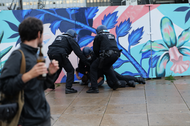 Eine Gruppe von Polizisten in schwarzer Einsatzkleidung steht neben einem Wandgemälde, einer hält eine Flasche, mit städtischen Strukturen im Hintergrund.