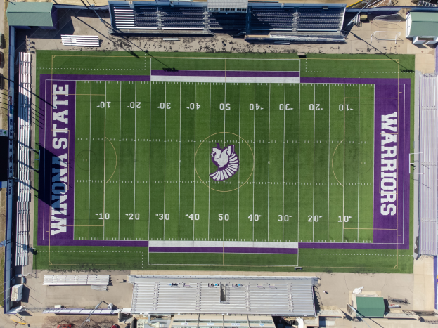 Eine Luftaufnahme eines Footballfelds mit violetten und weißen Markierungen, umgeben von Gebäuden, Fahrzeugen und Polen, die das Heimfeld der Footballmannschaft der Minnesota State University zeigt.