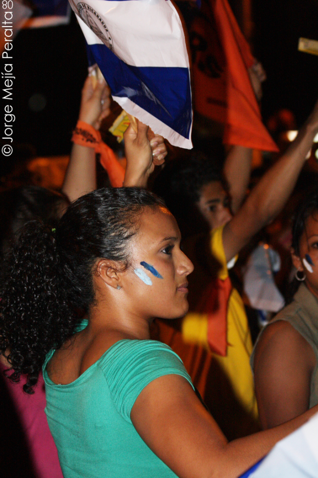 Eine Frau in einem grünen T-Shirt steht auf einer Demonstration und hält ein weißes Schild in der Hand, während eine Gruppe von Menschen hinter ihr steht.