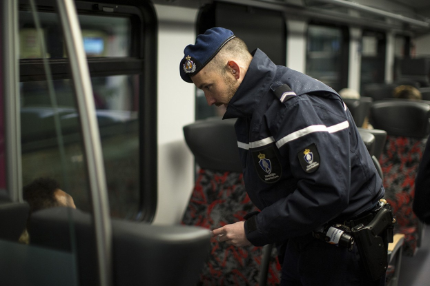 Ein Polizeibeamter in Uniform und Mütze steht in einem Zug und schaut auf sein Handy, umgeben von ein paar sitzenden Passagieren und einer weiteren Person auf der rechten Seite.