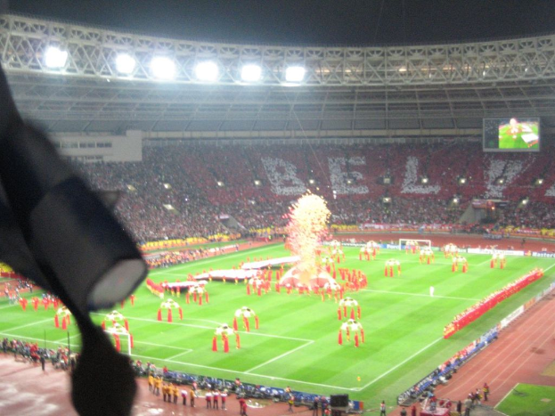 Ein Stadion mit Flutlicht, Eisengittern, Zuschauern, stehenden Personen, Werbetafeln, Anzeigetafeln, eine Überwachungskamera, Fahnen und Dekorationen.