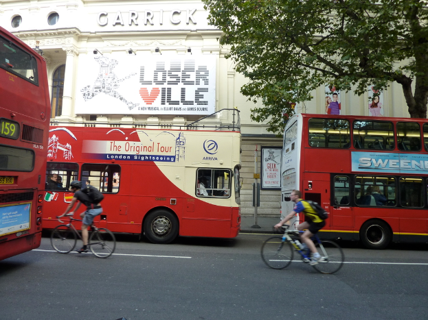 Mehrere Busse und Fahrradfahrer auf einer Straße, mit einem Haus, das Plakate zeigt, und einem Baum im Hintergrund.