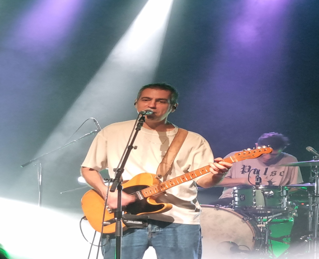 Ein Mann, der auf der Bühne eine E-Gitarre spielt und in ein Mikrofon singt, während ein anderer Musiker im Hintergrund Schlagzeug spielt.