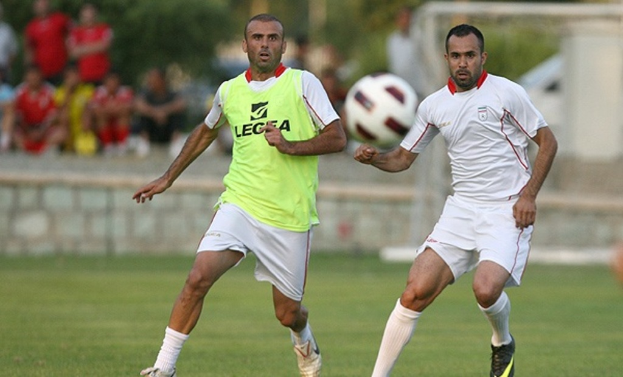 Zwei Spieler auf einem Rasenfeld mit einem Ball in der Luft, Zuschauer und Bäume im Hintergrund.